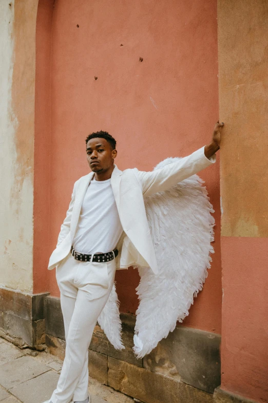 a man in a white suit leaning against a wall, an album cover, by Cosmo Alexander, pexels contest winner, renaissance, winged boy, miguel angel, wide open wings, androgynous person