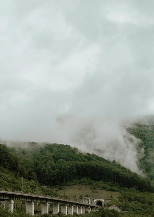 a train traveling down train tracks next to a lush green hillside, a picture, inspired by Elsa Bleda, pexels contest winner, romanticism, volumetric clouds and fog, panorama view, grey, low quality photo