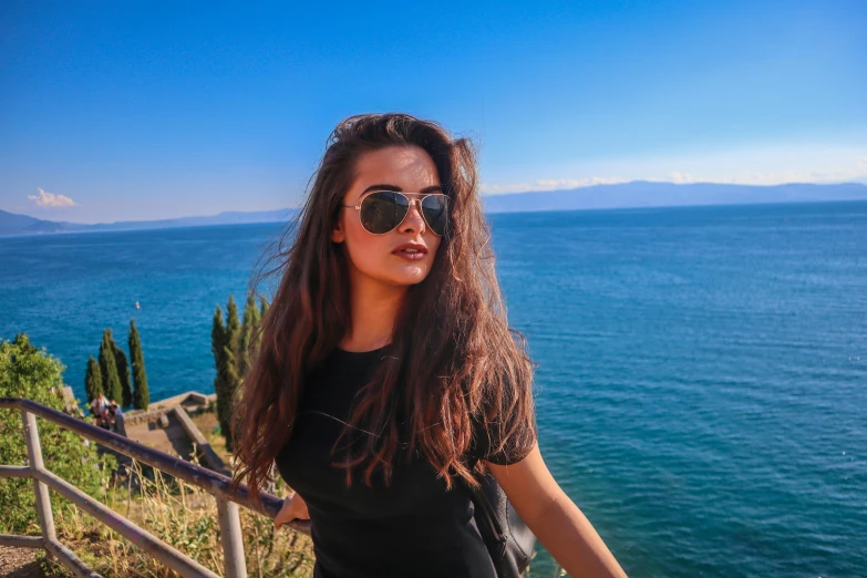 a woman standing in front of a body of water, fair olive skin, avatar image, wear ray - ban glass, sofya emelenko