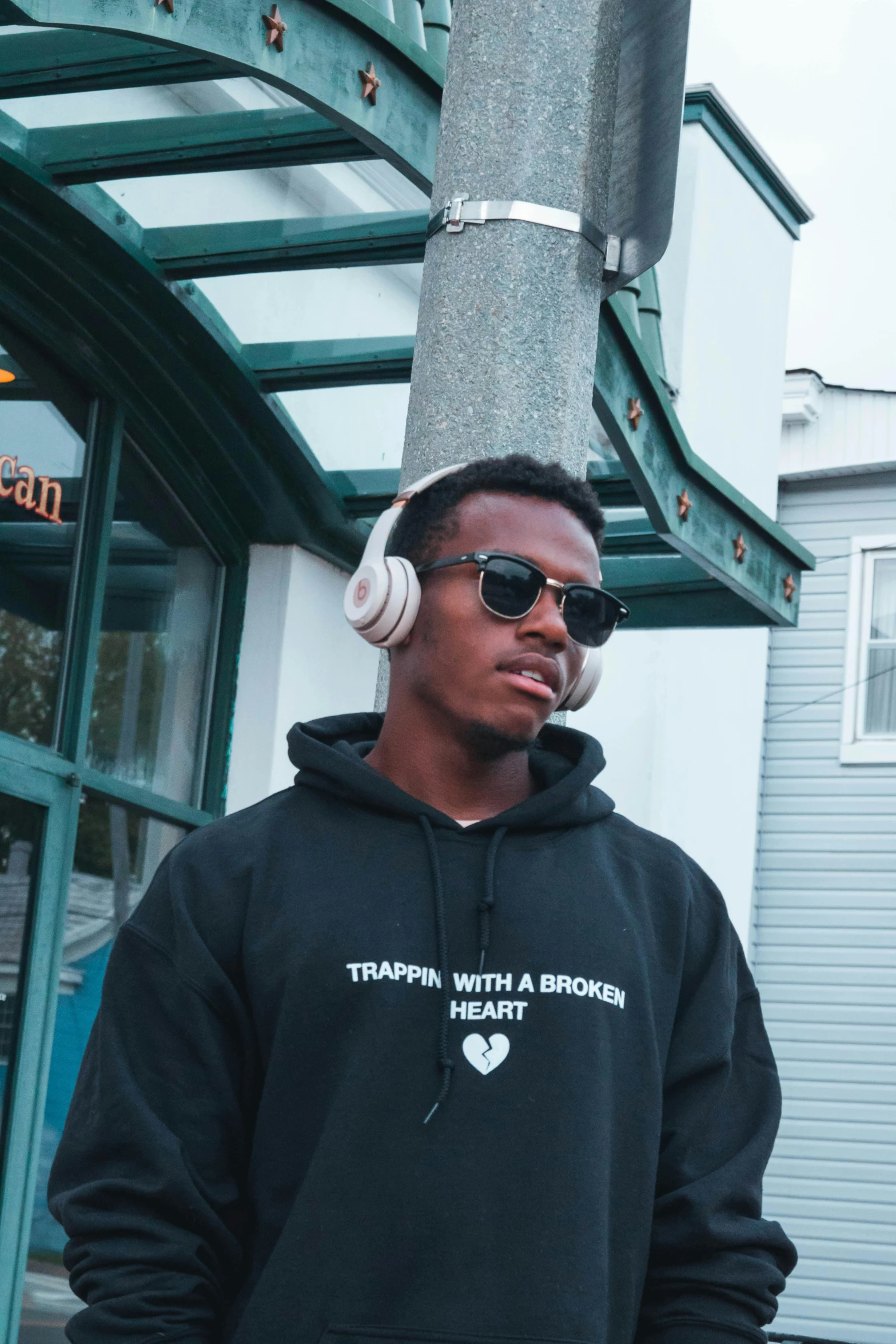 a man with headphones standing in front of a building, an album cover, inspired by Barthélemy Menn, trending on unsplash, hoodie, transgender, malcolm hart, wholesome