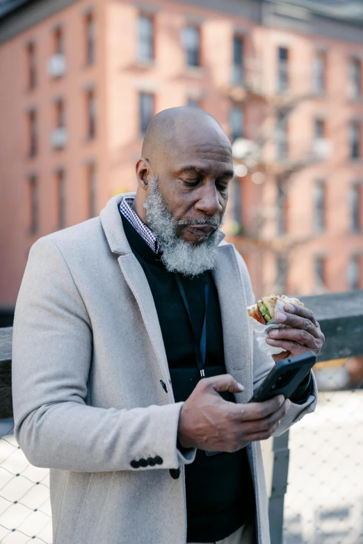a man standing next to a fence eating a sandwich, inspired by James E. Brewton, pexels contest winner, renaissance, bald head and white beard, he is holding a smartphone, harlem, people outside eating meals