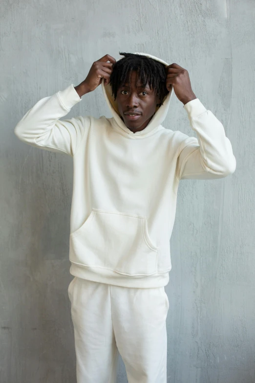 a man in a white hoodie standing in front of a gray wall, off - white collection, east african man with curly hair, wearing a hoodie and sweatpants, us