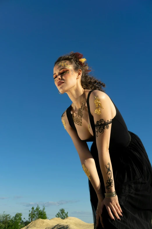 a woman in a black dress standing on a rock, an album cover, unsplash, renaissance, golden face tattoos, cloudless sky, gold bodypaint, body covered in floral tattoos