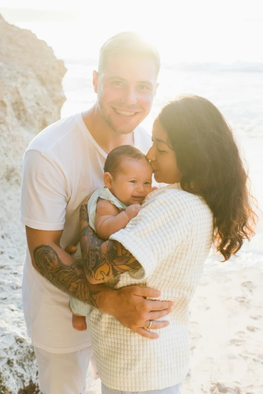 a man and woman holding a baby on the beach, with tattoos, radiant soft light, uploaded, white