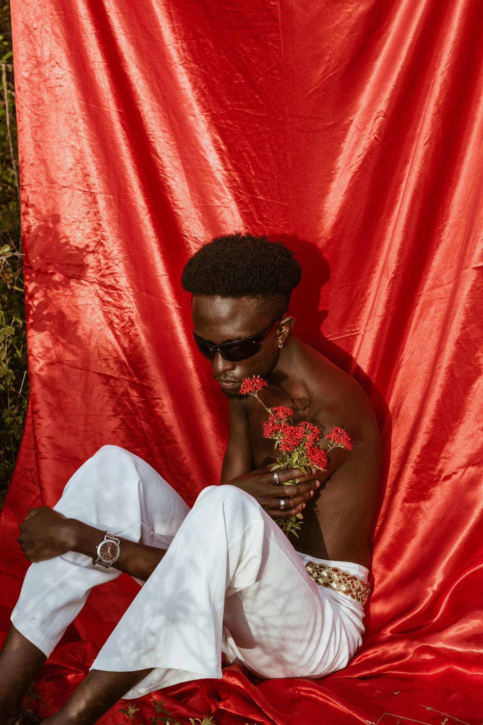 a man sitting on top of a red blanket, an album cover, inspired by David Bailly, trending on pexels, aestheticism, tall flowers, gold chains, red skin, delicate androgynous prince