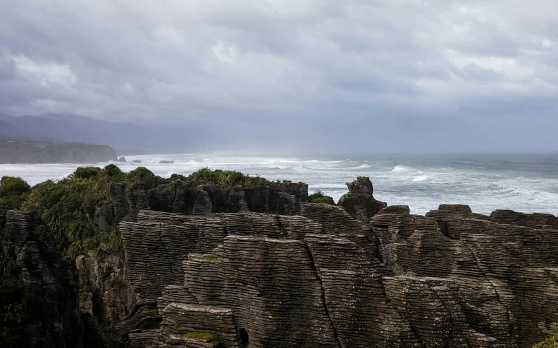 a man standing on top of a cliff next to the ocean, hurufiyya, chiseled formations, grey skies, kahikatea, stacked image