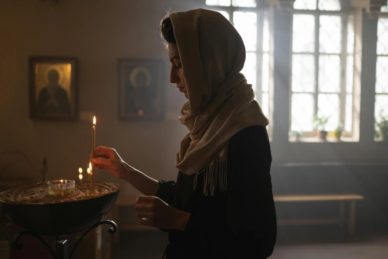 a woman lighting a candle in a dark room, head scarf, scene set in a church, perfectly lit. movie still, instagram post