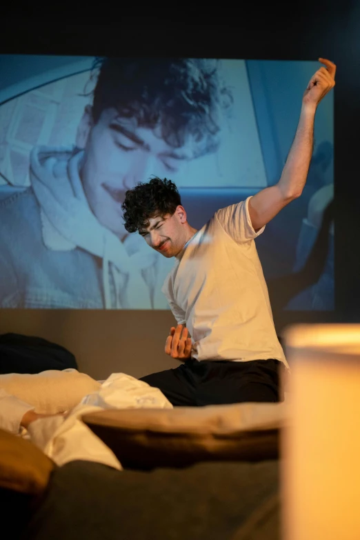 a man sitting on a bed in front of a projector screen, inspired by Nan Goldin, unsplash, happening, 3 actors on stage, declan mckenna, performing on stage, ( ( theatrical ) )