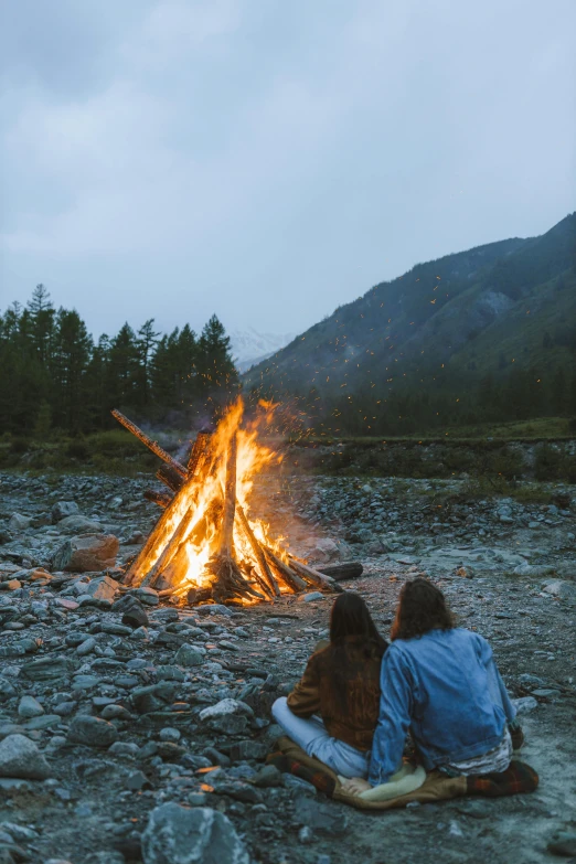 two people sitting in front of a campfire, a picture, unsplash contest winner, land art, beside the river, in mountains, grey, ryan mcginley