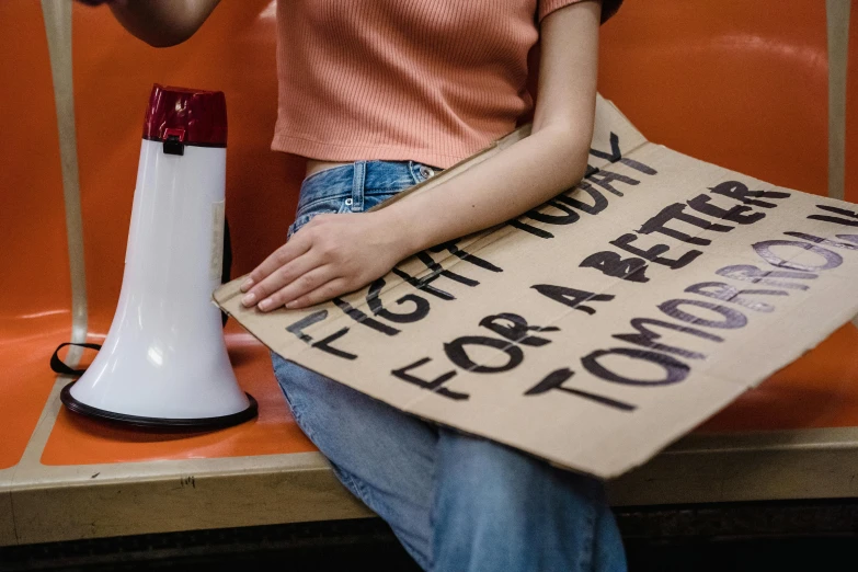 a woman sitting on a bench holding a sign, a poster, trending on pexels, graffiti, orange lamp, robotic prosthetic arm, protest, holding a tin can