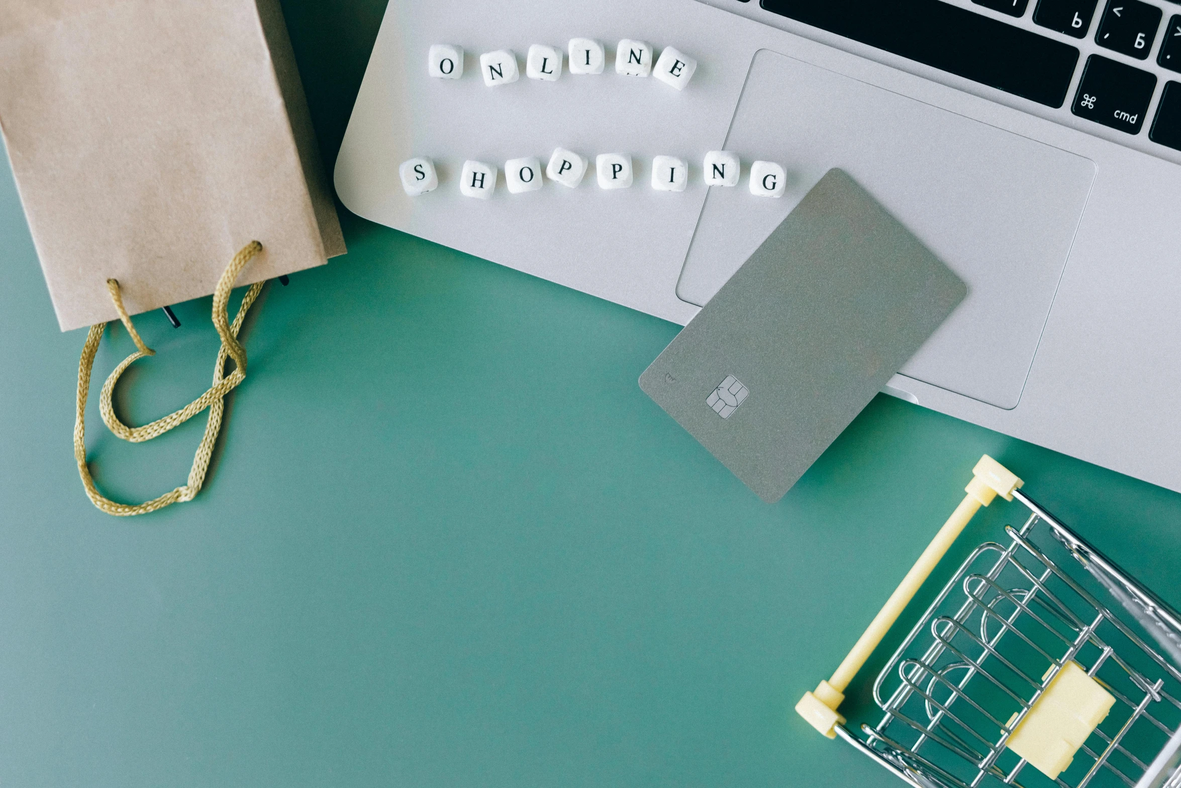 a laptop computer sitting on top of a desk next to a shopping cart, by Julia Pishtar, trending on pexels, happening, green letters, white and teal metallic accents, knolling, pair of keycards on table