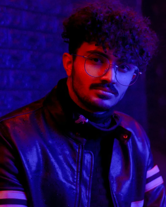 a man wearing glasses and a leather jacket, inspired by Max Dauthendey, trending on pexels, holography, curly haired, blacklight aesthetic, roshan, looking towards camera