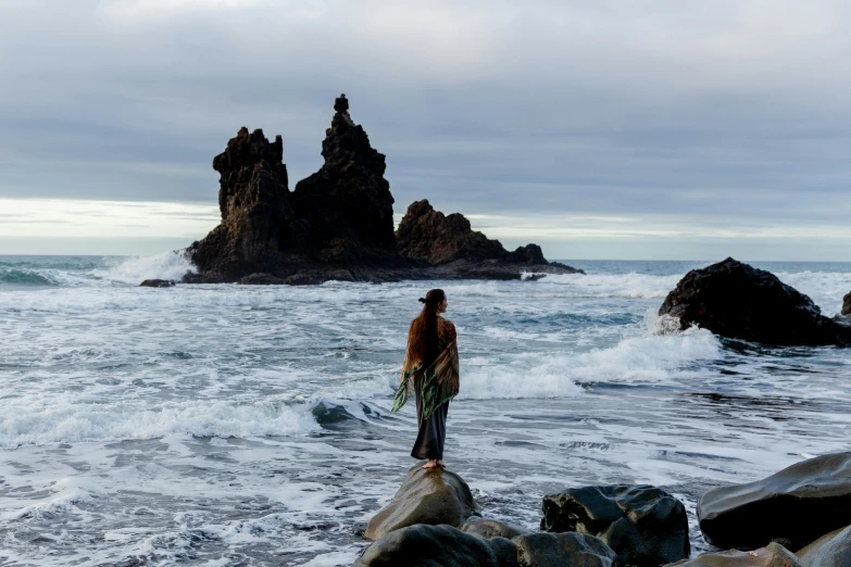 a man standing on top of a rock next to the ocean, by Terese Nielsen, unsplash contest winner, jason momoa, black sand, mermaids in distance, movie still 8 k