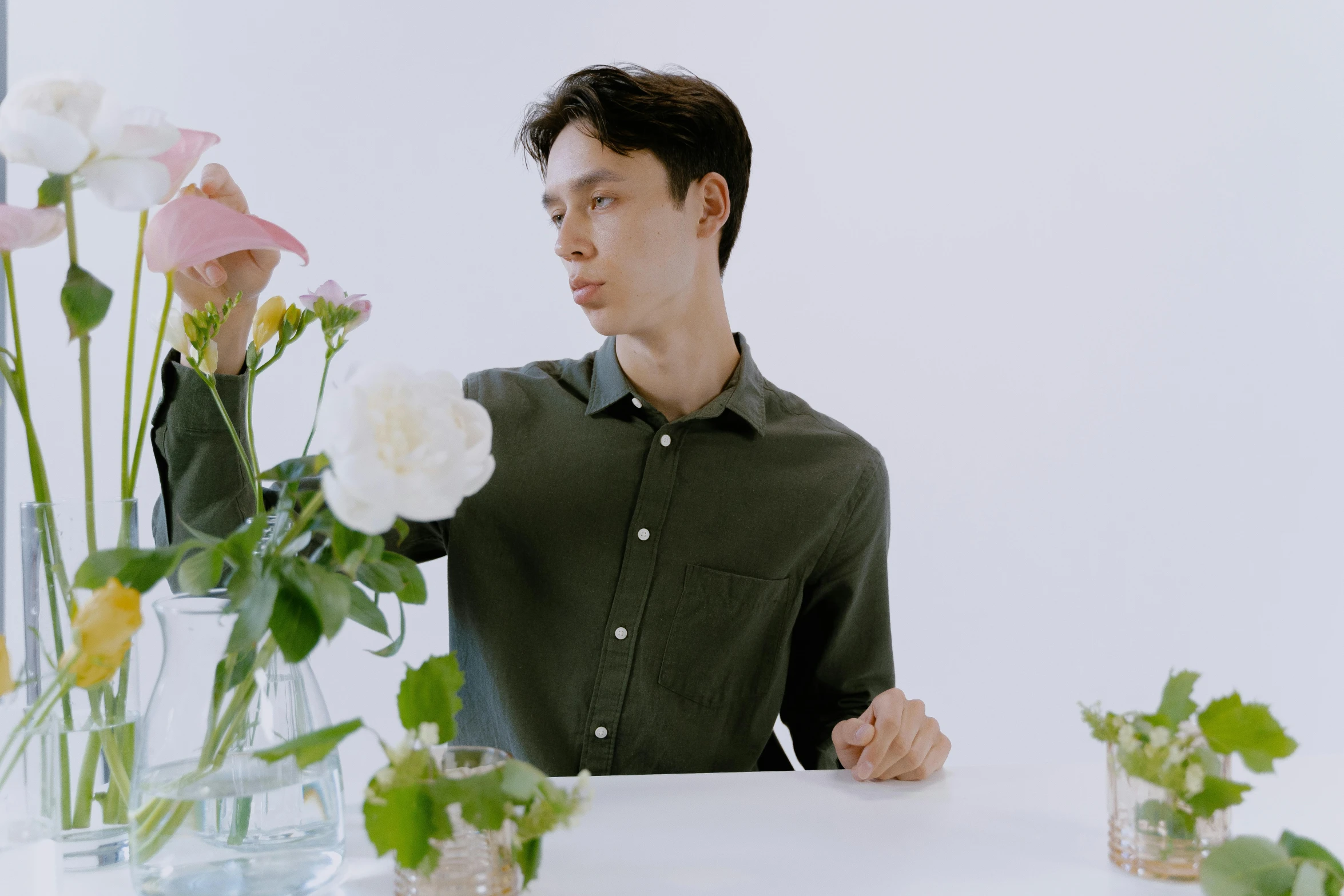 a man sitting at a table with flowers in vases, an album cover, inspired by Fei Danxu, in a dark green polo shirt, button up shirt, picking up a flower, well-groomed model