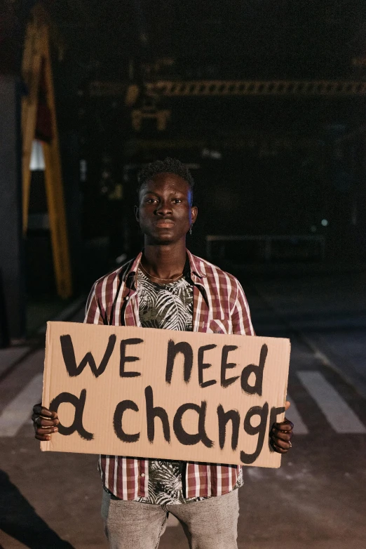 a man holding a sign that says we need change, an album cover, trending on unsplash, adut akech, people at night, mechanic, documentary still