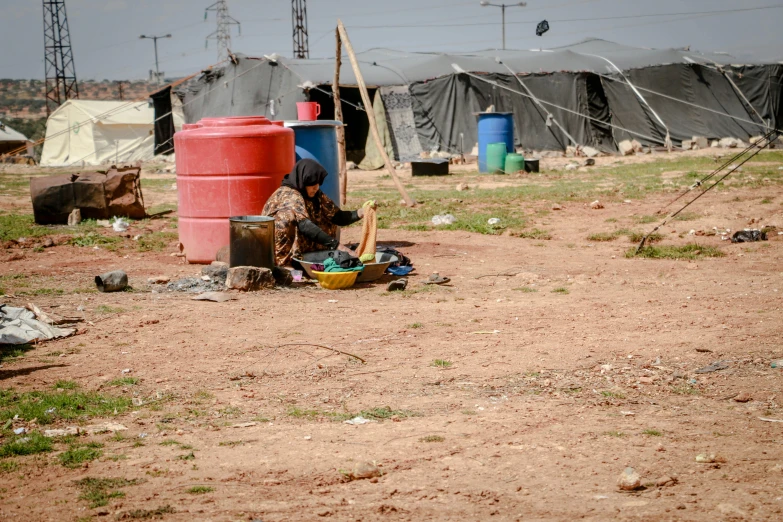 a woman sitting on the ground next to a red barrel, hurufiyya, barrel fires and tents, maintenance photo, 💣 💥💣 💥, a messy