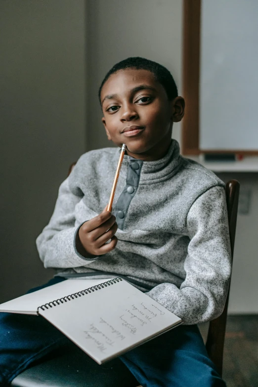 a young boy sitting in a chair with a pencil in his hand, pexels contest winner, ashcan school, black teenage boy, looking confident, boy with neutral face, holding notebook