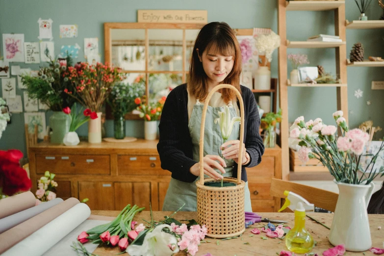 a woman arranging flowers in a basket on a table, inspired by Ruth Jên, pexels contest winner, arts and crafts movement, quirky shops, gif, louise zhang, in a short round glass vase