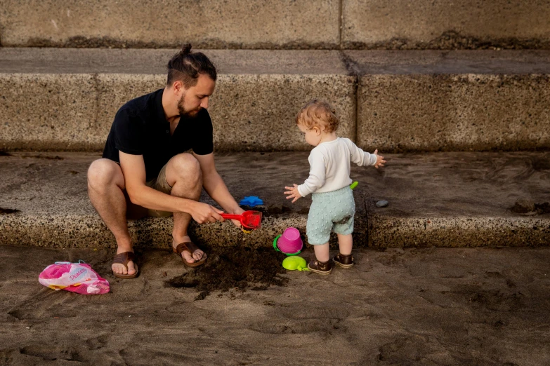 a man and a child playing in the sand, by Matt Stewart, pexels contest winner, in an urban setting, waterfals, toddler, low quality photo