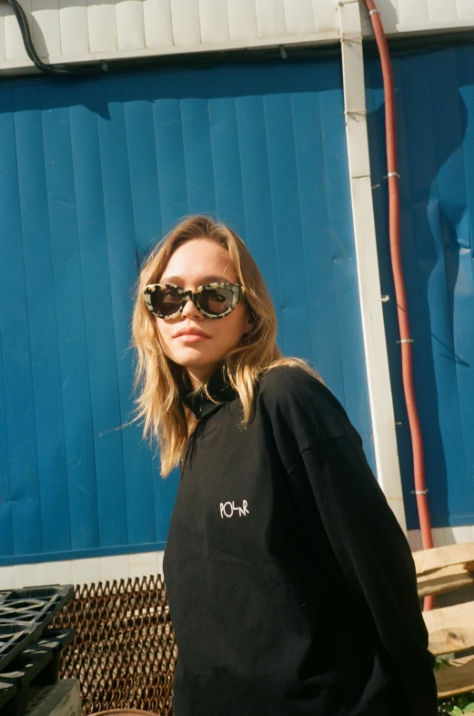 a woman standing in front of a blue building, an album cover, trending on unsplash, wearing a black sweater, in sun glasses, sydney sweeney, inhabited initials