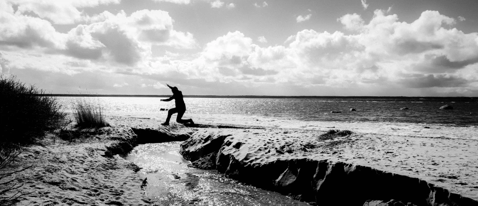 a man flying a kite on top of a sandy beach, a black and white photo, by Colijn de Coter, unsplash, figuration libre, leaping with arms up, near a jetty, flying mud, siluette