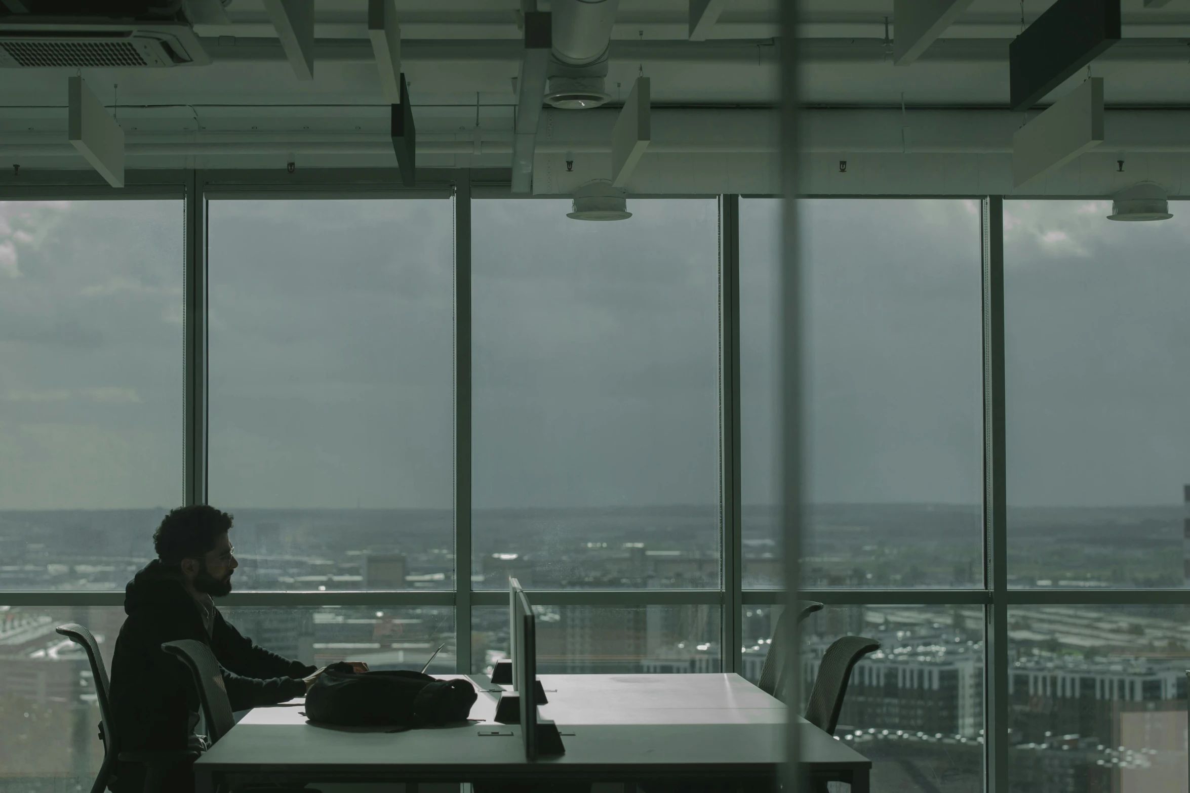 a man sitting at a table working on a laptop, by David Begbie, gloomy skies, **cinematic, glass ceilings, filmstill