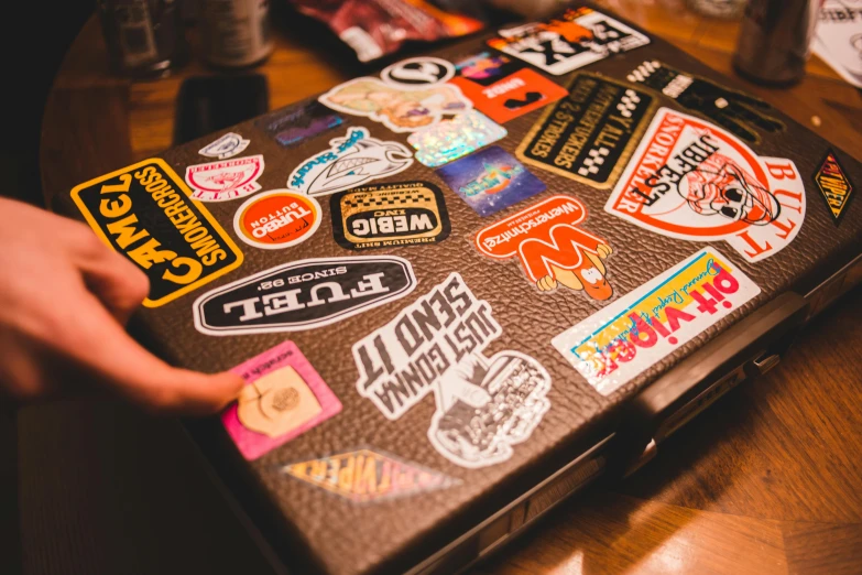 a person holding a piece of luggage covered in stickers, trending on pexels, patch logo design, laptops, grainy vintage, style of tony hawk pro skater