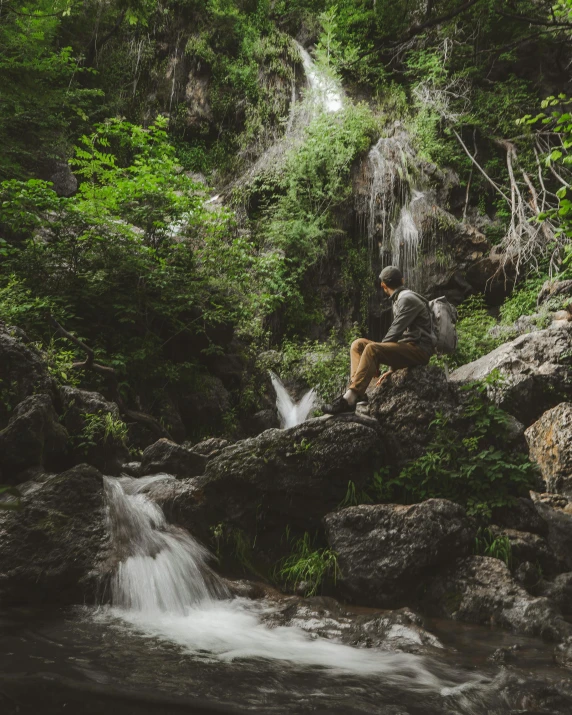 a man sitting on a rock in front of a waterfall, unsplash contest winner, verdant and lush and overgrown, al fresco, sitting in a tree, creeks