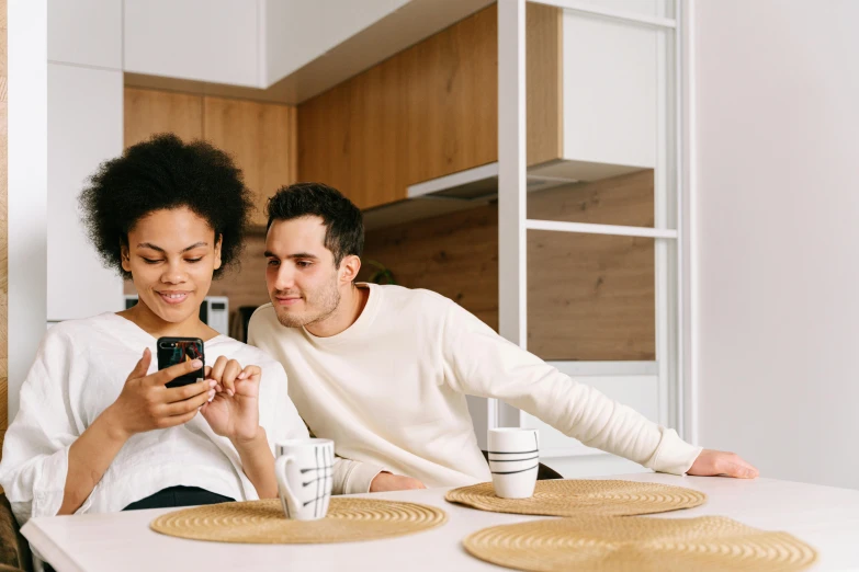 a man and woman sitting at a table looking at a cell phone, trending on pexels, happening, with a white mug, sassy pose, thumbnail, in small room