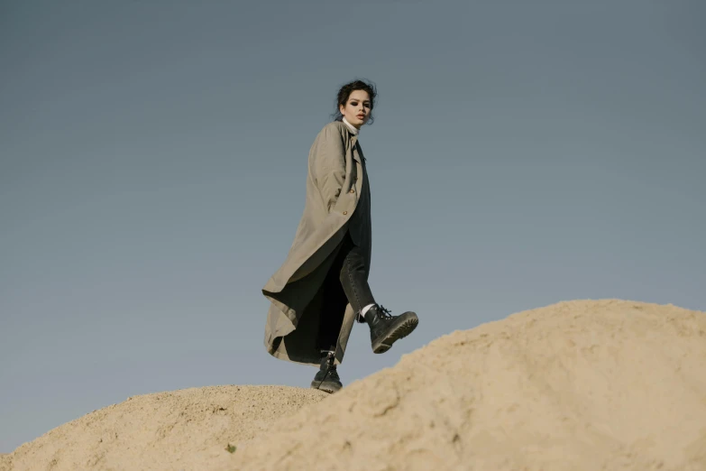 a man standing on top of a pile of sand, an album cover, unsplash, bauhaus, woman is in a trenchcoat, timothee chalamet, wearing tall combat boots, desert robe