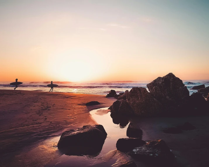 a couple of people standing on top of a sandy beach, during a sunset, rocks, surfing, lone person in the distance