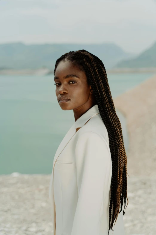 a woman standing in front of a body of water, brown skin. light makeup, long braided hair pulled back, wearing white suit, hammershøi