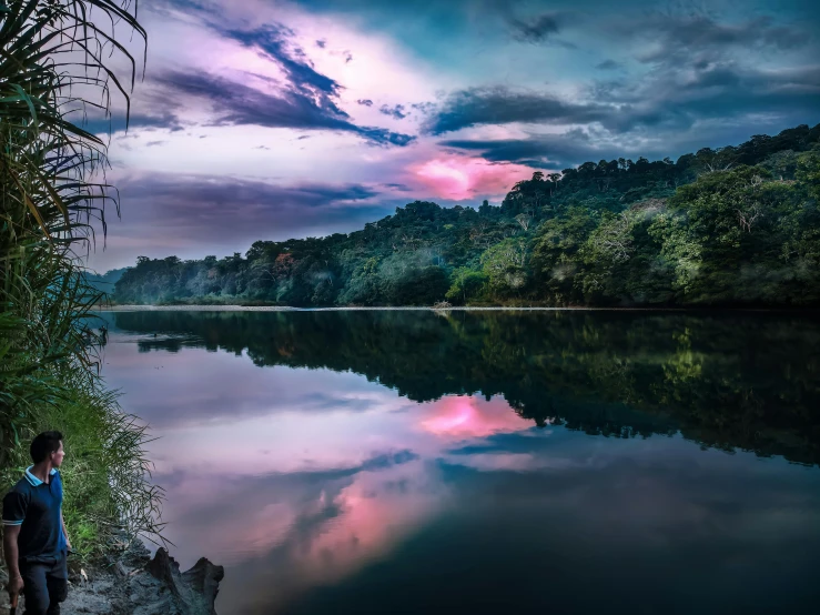 a man sitting on a rock next to a body of water, a matte painting, by Niklaus Manuel, pexels contest winner, sumatraism, pink reflections, scenic view of river, sri lanka, iridescence reflecting