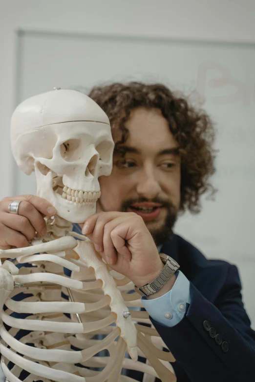 a man holding a model of a human skeleton, reddit, academic art, curls, still frame, teaching, looking towards camera