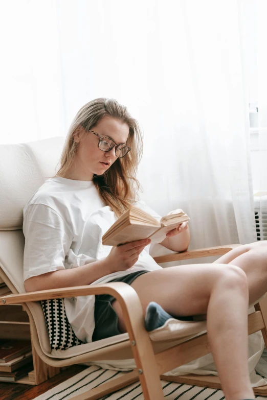 a woman sitting in a chair reading a book, pexels contest winner, wearing shorts and t shirt, wearing a light shirt, androgynous person, bare thighs