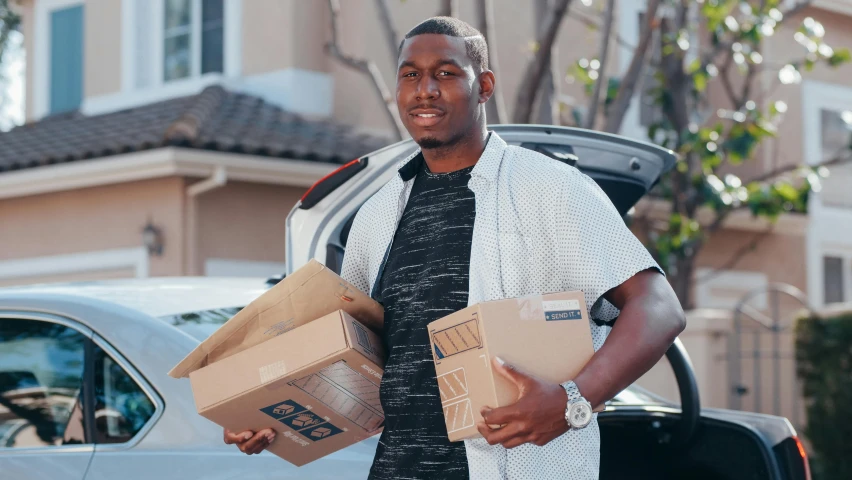 a man holding two boxes in front of a car, by Marshall Arisman, pexels contest winner, kayne west, bowater charlie and brom gerald, at home, professional sports style