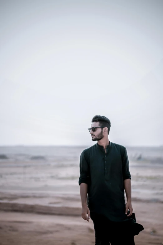 a man standing on a beach next to the ocean, a picture, by Ismail Acar, minimalism, wearing black glasses, standing in a barren field, wearing a kurta, focused stare