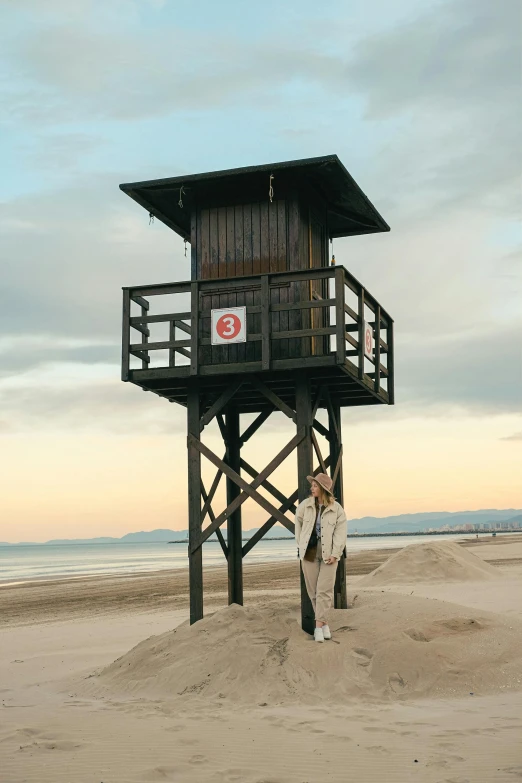 a woman standing on top of a sandy beach next to a lifeguard tower, ana de la reguera portrait, wooden toilets, evan lee, high towers