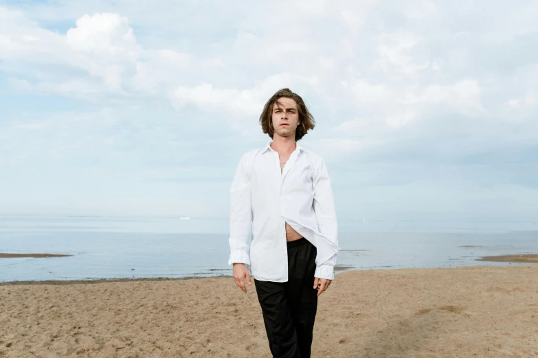 a man standing on top of a sandy beach, an album cover, by Attila Meszlenyi, unsplash, wearing a white button up shirt, timothee chalamet, andrea savchenko, mid long hair