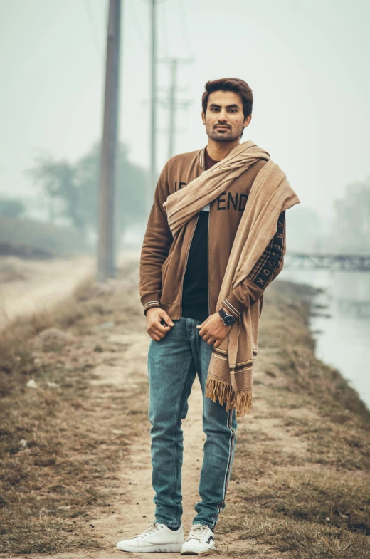 a man standing on a dirt road next to a body of water, a picture, inspired by Sunil Das, wearing a scarf, 4k fashion shoot, he is wearing a brown sweater, jeans and t shirt