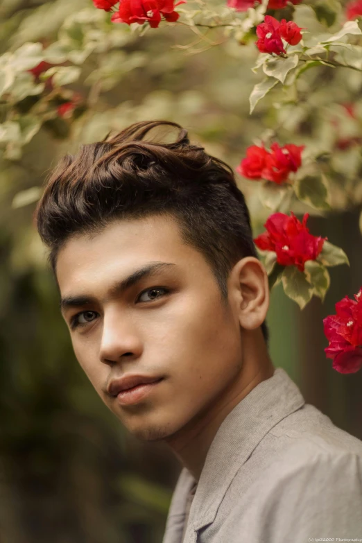 a young man standing in front of a bush of red flowers, unsplash, sumatraism, hair styled in a bun, square masculine facial features, hair : long brown, 5 0 0 px models