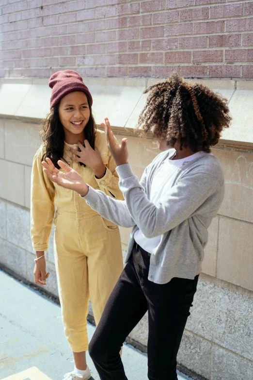 a couple of women standing next to each other on a sidewalk, trending on pexels, happening, greeting hand on head, yellow clothes, black teenage girl, dancing with each other