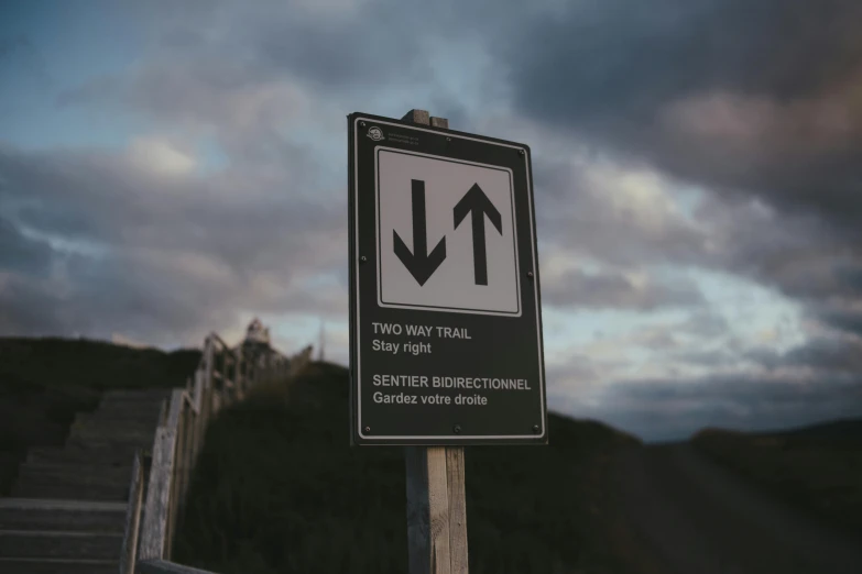 a black and white sign sitting on the side of a road, by Adam Szentpétery, unsplash, on a cliff, with two arrows, in the evening, zig zag