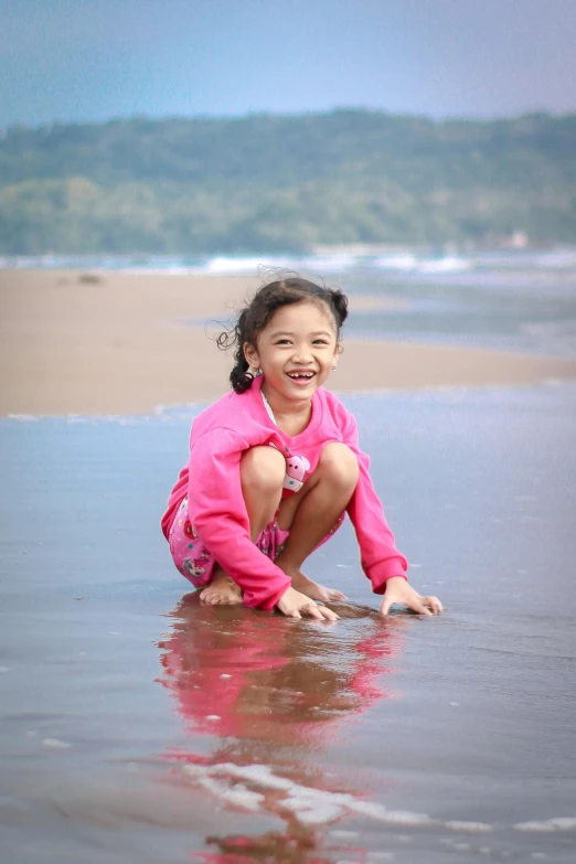 a little girl sitting on top of a sandy beach, in the water, while smiling for a photograph, kakar cheung, pink