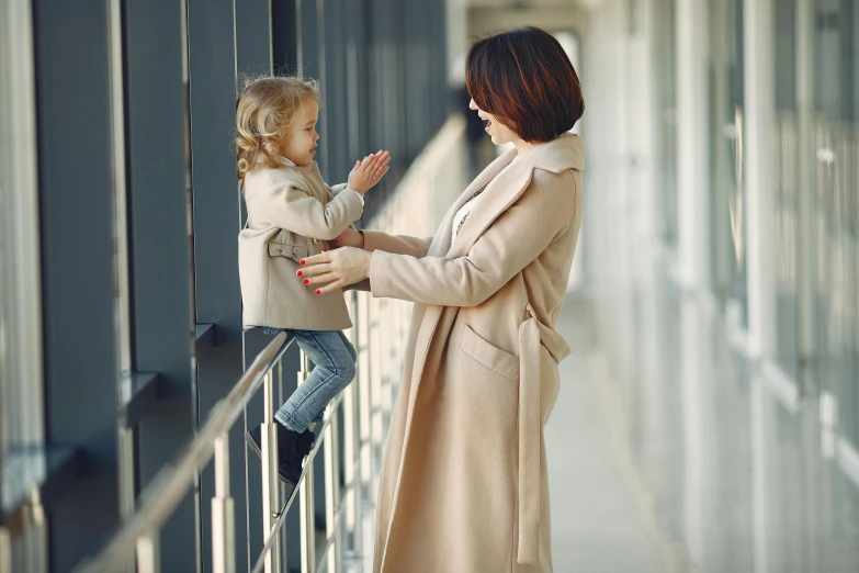 a woman standing next to a little girl on a balcony, pexels contest winner, symbolism, light brown coat, airport, standing on a shelf, holding hand