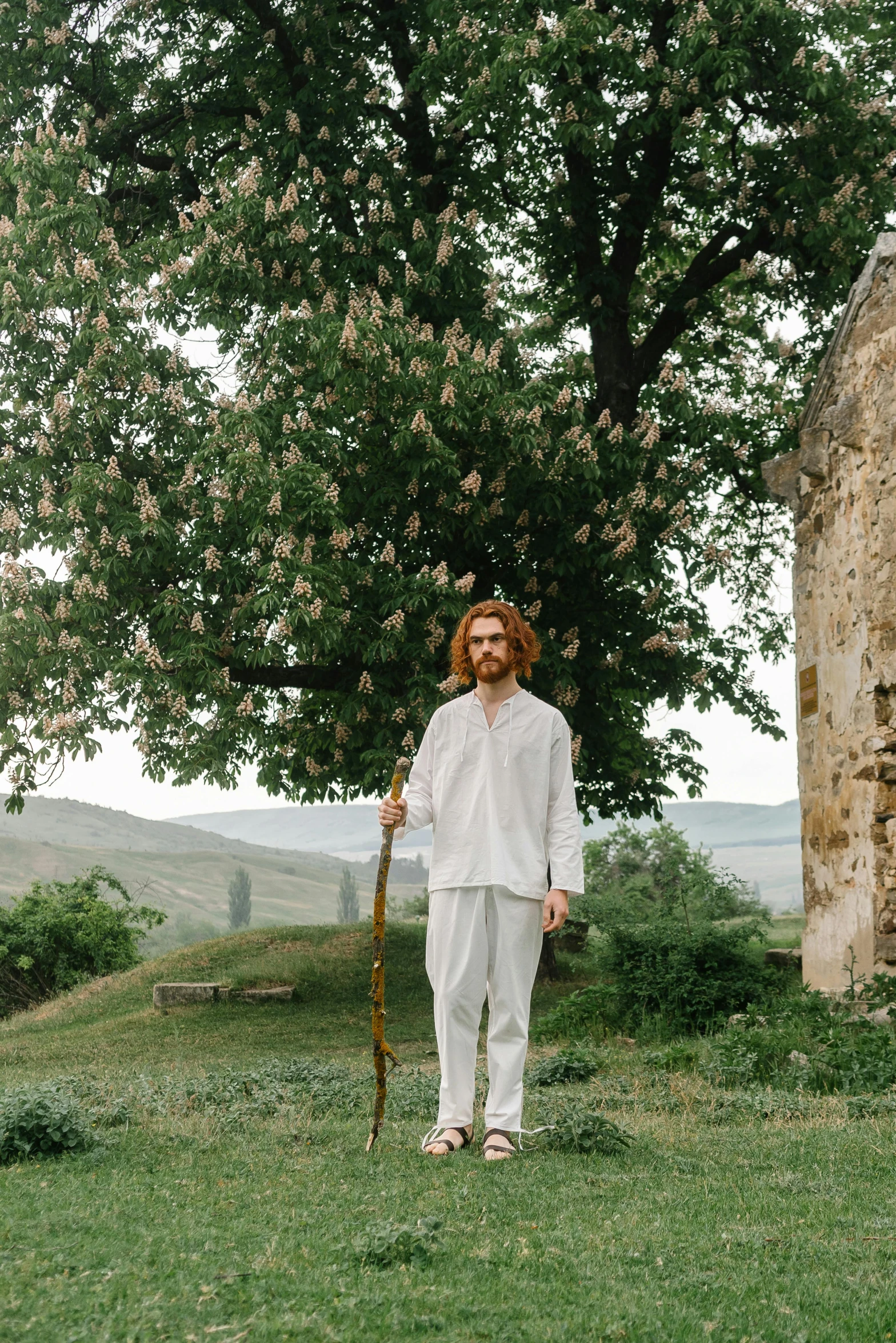 a man that is standing in the grass with a stick, an album cover, inspired by Apollinary Vasnetsov, unsplash, renaissance, wearing white pajamas, clothed in ancient street wear, hills, hozier