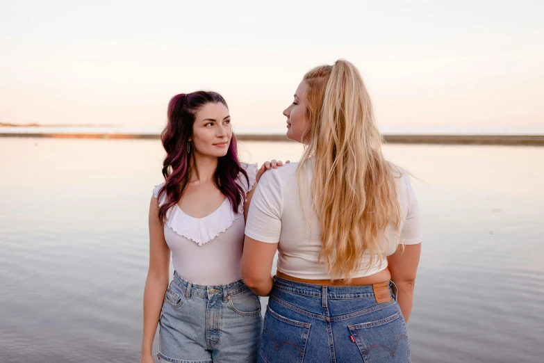 two women standing next to each other near a body of water, trending on pexels, one blonde and one brunette, plain background, lesbian, calmly conversing 8k