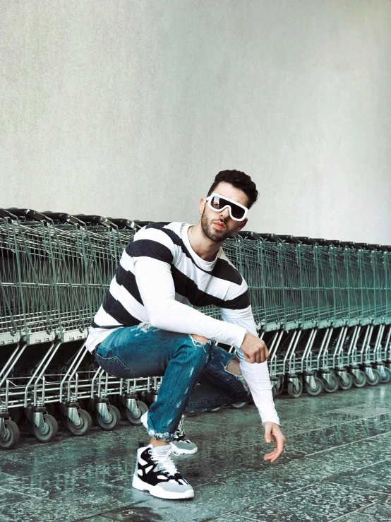 a man squatting in front of a row of shopping carts, an album cover, inspired by Maximilian Cercha, trending on pexels, photorealism, striped sweater, wearing cool sunglasses, confident pose, young greek man