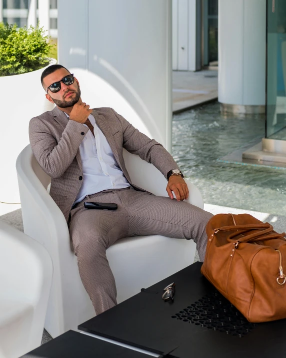 a man sitting on a white chair with a brown bag, inspired by Károly Brocky, trending on instagram, wearing tight suit, miami beach, lgbtq, platinum attire