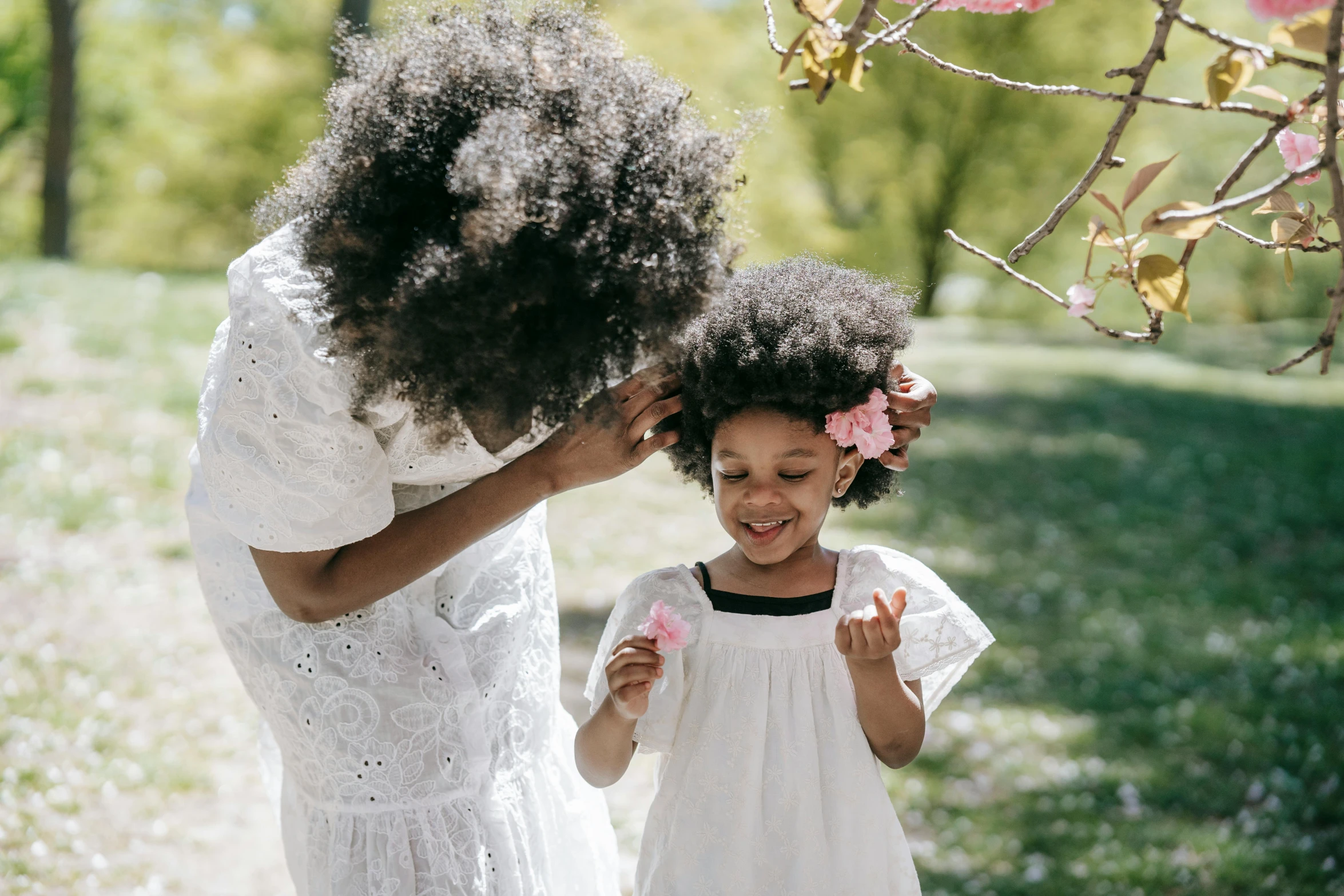 a woman standing next to a little girl in a white dress, by Lily Delissa Joseph, pexels contest winner, symbolism, afro comb, sakura season, with curls, manuka